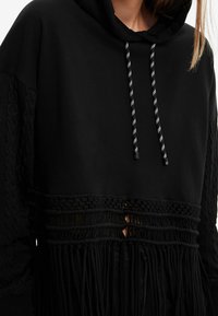 Sudadera negra con acentos de punto en las mangas, un diseño de macramé en la cintura y cordones con un patrón a rayas.