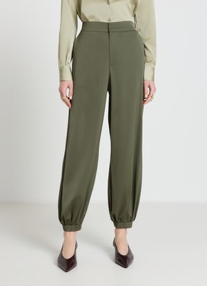 Frau trägt olivgrüne, hoch taillierte Hosen mit elastischen Bündchen, kombiniert mit einem hellbeigen Hemd mit Knöpfen und dunklen, spitzen Absätzen.