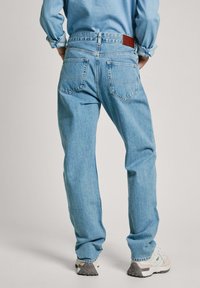 Gai zila denim bikses ar brīvu piegriezumu, kurām ir divi aizmugurējie kabati, ādas jostas ielāps un sarullēti piedurknes, valkātas ar gaišiem sporta apaviem.