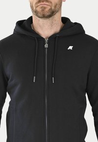 Sweat à capuche noir zippé en tissu côtelé. Il dispose d'une fermeture éclair argent ée et de cordons de serrage, avec un petit logo sur le côté supérieur gauche.