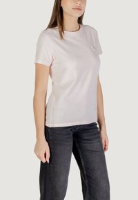 T-shirt in cotone rosa con colletto rotondo e maniche corte, dotata di un piccolo logo triangolare sul lato sinistro, abbinata a jeans scuri.