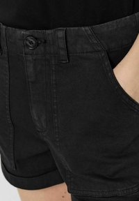 Shorts en denim noir avec fermeture à bouton et ourlet retroussé, portés avec un haut noir et une main glissée dans la poche droite.