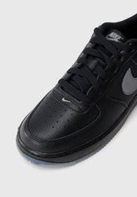 Crne kožne tenisice s mrežastim detaljima, sivi Nike logo, okruglice za vezivanje i teksturirana crna gumena potplat. Perforirana zona za prste.