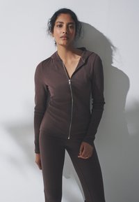 Felpa con zip marrone a coste con design aderente e cappuccio, abbinata a leggings marroni aderenti coordinati. Tessuto liscio, dettaglio di zip metallica.