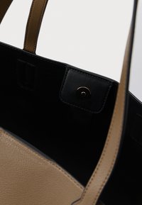 Geantă tote din piele deschisă, cu un finisaj texturat, interior negru, curele contrastante și un sistem de închidere metalic rotund în partea de sus.