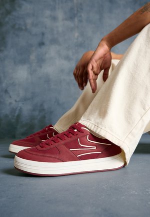 Persoon die gebroken witte broek en rode sneakers met witte zolen draagt, zittend op een grijze vloer met handen die rusten nabij de knieën.