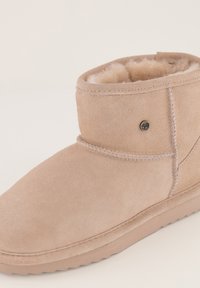 Beige suède enkellaars met schapenvachtvoering, ronde neus en platformzool. Bevat stiksels en een klein cirkelvormig logootje.