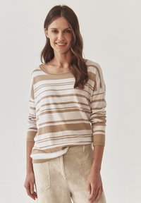 Pull rayé beige et blanc avec un col large et des manches longues, fabriqué en matériau doux. Porté avec un pantalon beige clair.