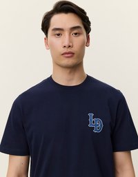 Tumši zils kokvilnas T krekls ar kontrastējošu gaiši zilu logotipu kreisajā krūtī, apaļu kakla izgriezumu un īsām piedurknēm. Gluda tekstūra.