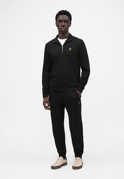 Homme portant un sweat-shirt noir à fermeture éclair quart de zip et un pantalon de jogging noir assorti avec des baskets beiges, debout devant un fond blanc uni.