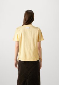Gelbes Baumwoll-T-Shirt mit kurzen Ärmeln, rundem Ausschnitt und einem Etikett auf der Rückseite. Kombiniert mit einem dunkelbraunen Rock.