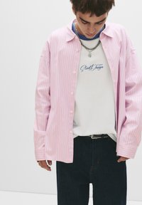 Cămașă roz deschis cu dungi, purtată deschis peste un tricou alb cu text albastru, asortată cu pantaloni din denim închis și o curea cu ținte.