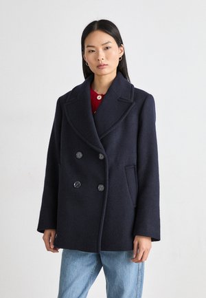 Marineblauer Woll-Doppelreiher mit großen Revers, zwei Knöpfen und Seitentaschen, kombiniert mit einem roten Oberteil und hellblauen Jeans.