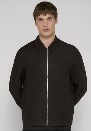 Young man with short brown hair wearing a black zip-up jacket with collar and chest pocket, standing against a plain background.