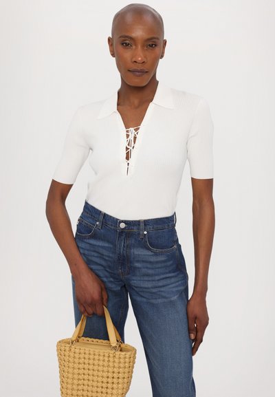 Camiseta de polo blanca con relieve y mangas cortas, con un escote repleto de cordones; combinada con jeans de mezclilla azules y un bolso amarillo de mimbre.