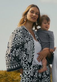 Schwarz-weiße bestickte Kimono-Jacke mit floralen Mustern, über einem weißen Oberteil getragen, kombiniert mit passenden Hosen. Baby in grauer Kleidung.