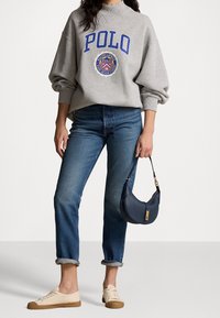 Felpa grigia con logo "POLO" blu, jeans blu, sneakers color crema e una borsa a tracolla blu scuro con una finitura dorata.