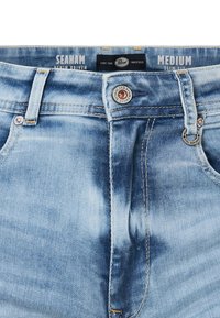 Jeans de mezclilla azul claro con un corte ajustado, bolsillos delanteros y detalles metálicos en los botones. La tela muestra una textura desgastada con detalles de desvanecimiento.