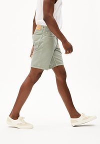 Lichtgroene katoenen shorts met twee achterzakken en een tan leren label; gedragen met witte sneakers met beige accenten.