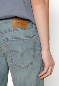 Ljusblå denimjeans med en blekt finish, utrustad med en läderpatch och V-stickning på bakfickorna. Bärs med en grå skjorta.