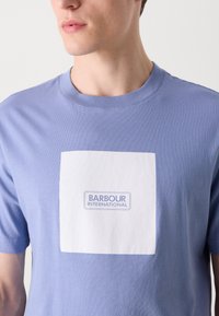 Camiseta de algodão azul claro com um retângulo branco com o texto azul "BARBOUR INTERNATIONAL" centrado no peito.