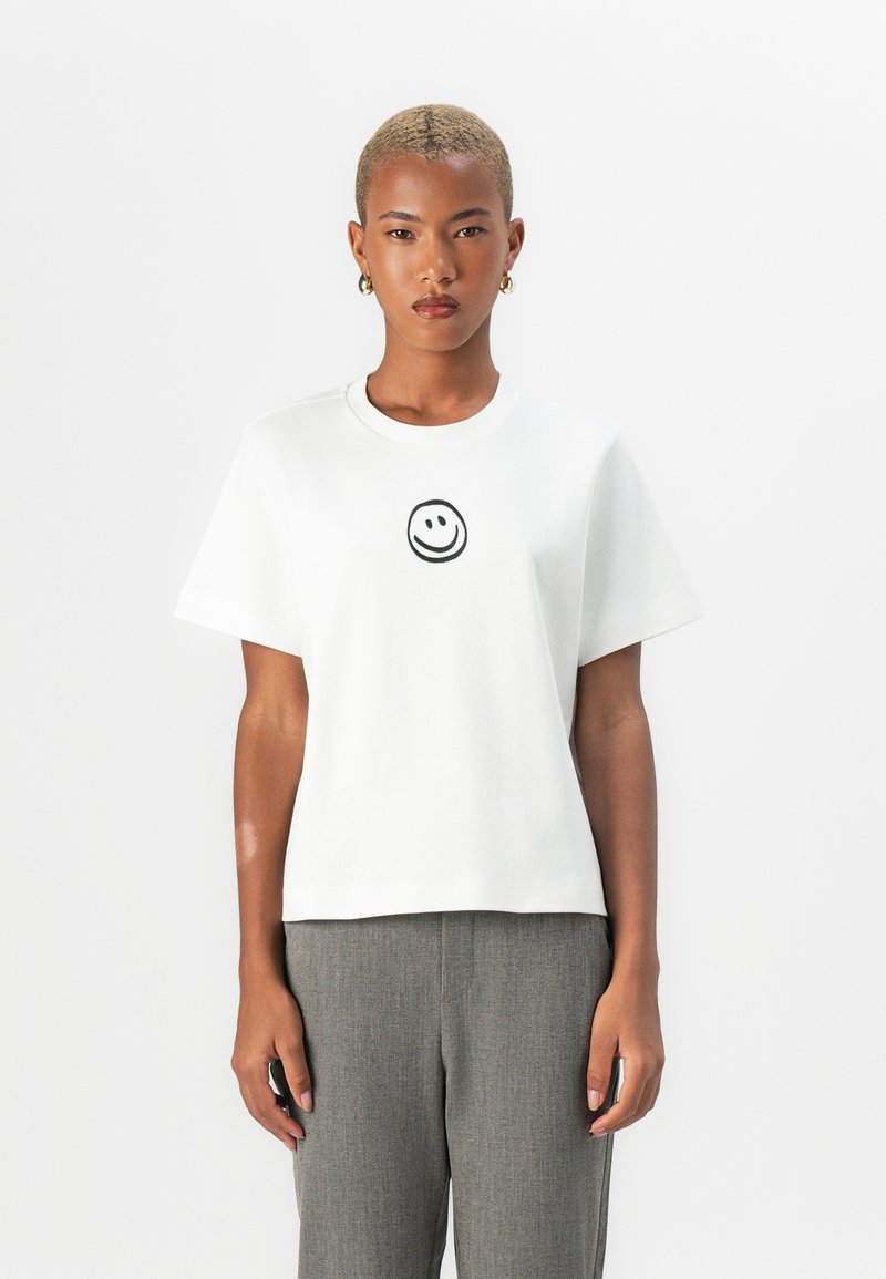 White cotton t-shirt featuring a black smiley face graphic at the center, short sleeves, a round neckline, and a relaxed fit.