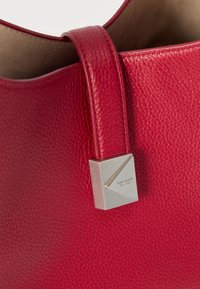 Κόκκινο λουρί τσάντας από δέρμα με υφή και ασημένια τετράγωνη αγκράφα χαραγμένη με την επιγραφή «kate spade NEW YORK» στο πλάι της τσάντας.