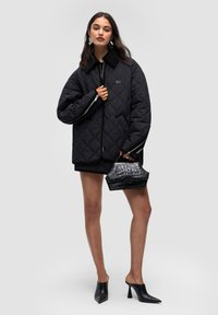 Giacca a vento nera trapuntata con colletto, zip argentate e logo ricamato. Il modello tiene in mano una piccola borsa nera in pelle di coccodrillo.