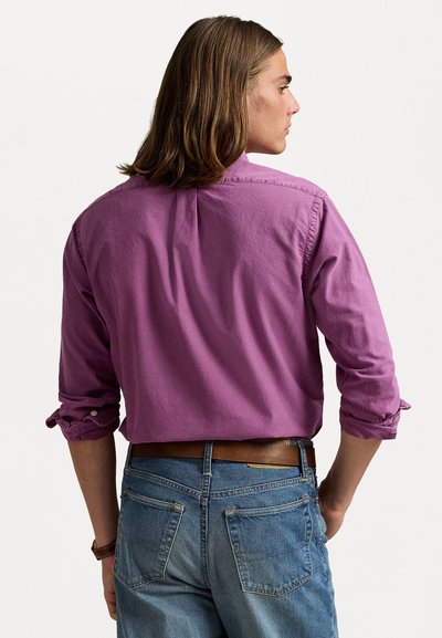 Långärmad lila skjorta, mjukt tyg, avslappnad passform, uppvikta ärmar, detaljer med bakställd yoke, kombinerad med ljusblå jeans och ett brunt bälte.