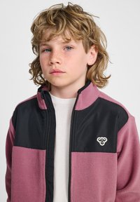 Fleecejack met een zwarte bovenkant en roze onderkant, voorzien van een ritssluiting aan de voorkant, een opstaande kraag en een klein embleem op de borst.