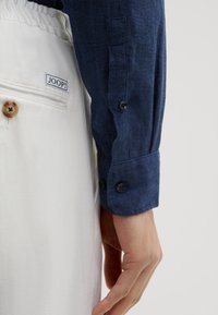 Navyblaues Leinenhemd mit langen Ärmeln und Knopfmanschetten, kombiniert mit weißen Hosen, die einen Knopf und ein Markenlabel aufweisen. Sichtbare Strukturdetails des Stoffes.