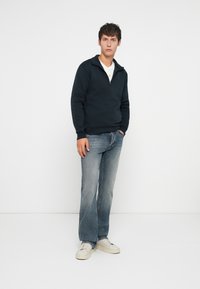 Pull marin à moitié zipper, assorti à un jean bleu clair et des baskets blanc cassé, sur un mannequin sur fond blanc.
