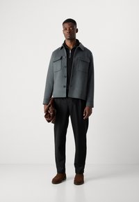 Gray wool jacket with button closure and two chest pockets, black zip-up top, black trousers, brown boots, holding a brown leather bag.