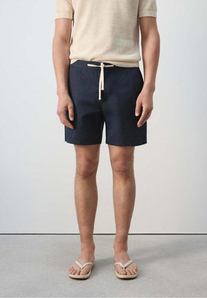 Hombre de pie con camiseta de manga corta beige, pantalones cortos azul marino con cordón y chanclas beige, delante de un fondo claro y liso.