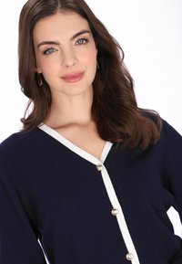 Laivastonsininen neuletakki valkoisilla resoreilla, V-kaula-aukolla ja kultaisen sävyn napeilla. Pehmeä rakenne ja pitkät hihat, painottaen klassista muotoilua.