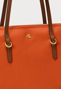 Πορτοκαλί καμβάς τσάντα tote με καφέ δερμάτινες λαβές, χρυσά αξεσουάρ και μια χρυσή λογότυπο λεπτομέρεια στην μπροστινή πλευρά. Διαθέτει κλείσιμο με φερμουάρ.