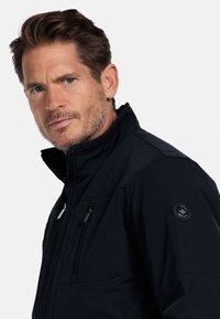 Donkerblauwe softshelljack met een hoge kraag, een ritssluiting aan de voorkant en een ritszak op de borst. Met een ronde logo op de mouw.