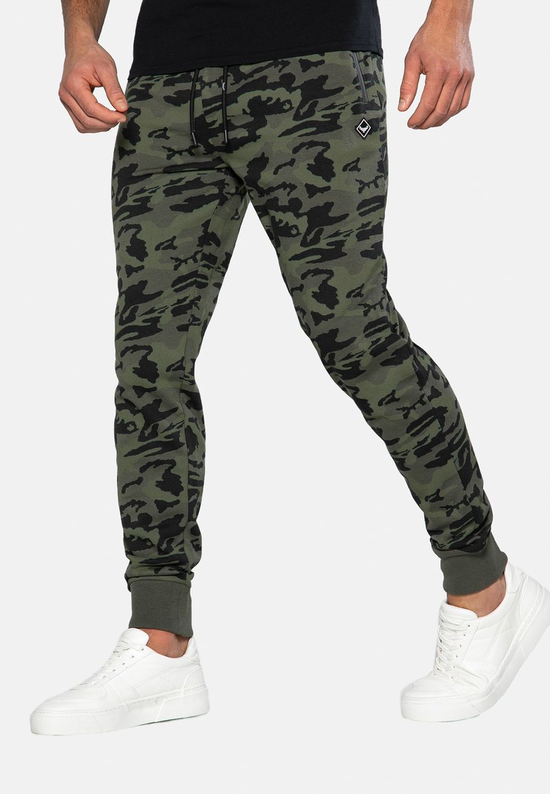 Tarnfarbene Joggerhosen in Grüntönen und Schwarz, aus weichem Stoff mit schmal zulaufendem Schnitt und gerippten Bündchen. Kombiniert mit weißen Sneakers.