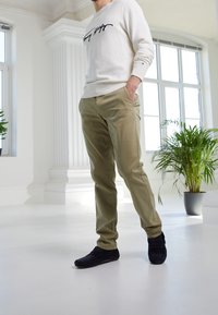 Beige Chinos mit lockerer Passform und Seitentaschen, kombiniert mit einem hellen Creme-Sweatshirt mit dunklem Schriftzug und gerippten Bündchen, getragen mit dunklen Loafers.