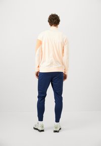 Light peach athletic jacket with long sleeves, paired with navy fitted pants and light-colored sneakers. Smooth fabric with contrasting color blocks.