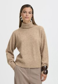 Maglione beige con collo alto, dalla vestibilità comoda e orlo a costine, caratterizzato da un hardware unico di accentuazione marrone scuro sulle maniche. Texture morbida.