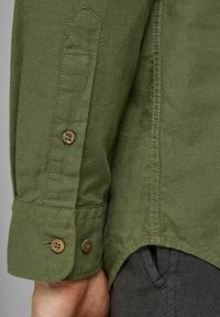 Lähikuva vihreästä pitkähihaisesta paidasta, jossa on kaksi ruskeaa nappia, yksityiskohtaista ompelua ja osa kädestä sekä tummat kangashousut näkyvissä.