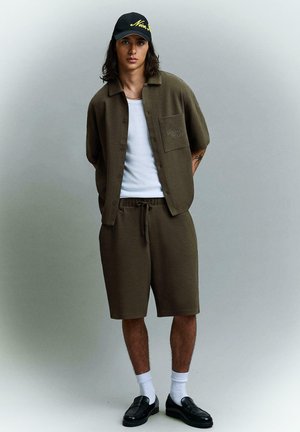 Fiatal férfi áll egy egyszínű háttér előtt, olíva zöld rövid ujjú ingben és rövidnadrágban, fehér trikóban, fekete sapkában, fehér zokniban és fekete mokaszinkban.