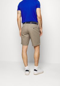 Hombre con camiseta azul, pantalones cortos beiges con un cinturón negro, calcetines blancos y zapatillas blancas, de pie de espaldas sobre un fondo blanco liso.
