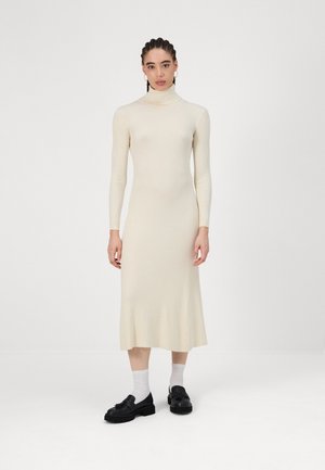 Kermavin ribbed midi-mekko pitkillä hihoilla ja korkealla kauluksella, jossa on vartalonmyötäinen yläosa, joka levenee helmaa kohti. Yhdistetty mustien kenkien kanssa.