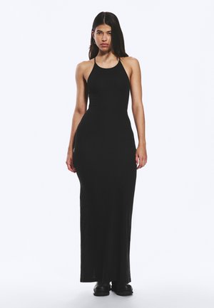 Frau mit langem, dunklem Haar, die ein ärmelloses, bodenlanges schwarzes Kleid und schwarze Schuhe trägt, vor einem schlichten weißen Hintergrund steht.