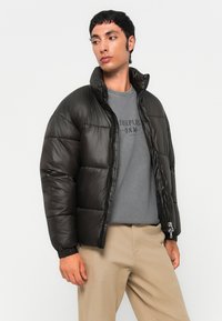Giacca nera imbottita con colletto alto e chiusura con zip. Texture liscia, polsini elastici, indossata sopra un maglione grigio e pantaloni beige.