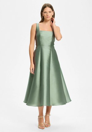 Femme portant une robe midi en satin vert sauge sans manches avec un décolleté carré et des talons dorés à brides, debout sur un fond blanc uni.