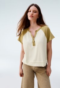 Short-sleeve top in light cream with olive green shoulder accents, featuring a buttoned neckline and a relaxed fit. Textured fabric, casual style.