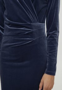 Robe en velours bleu marine à manches longues, avec tissu froncé et silhouette ajustée. La texture est lisse et brillante.
